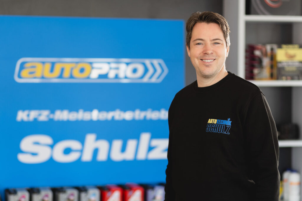 Ein Mitarbeiter von Autotechnik Schulz steht vor einem Schild mit dem Logo und Schriftzug des KFZ-Meisterbetriebs.